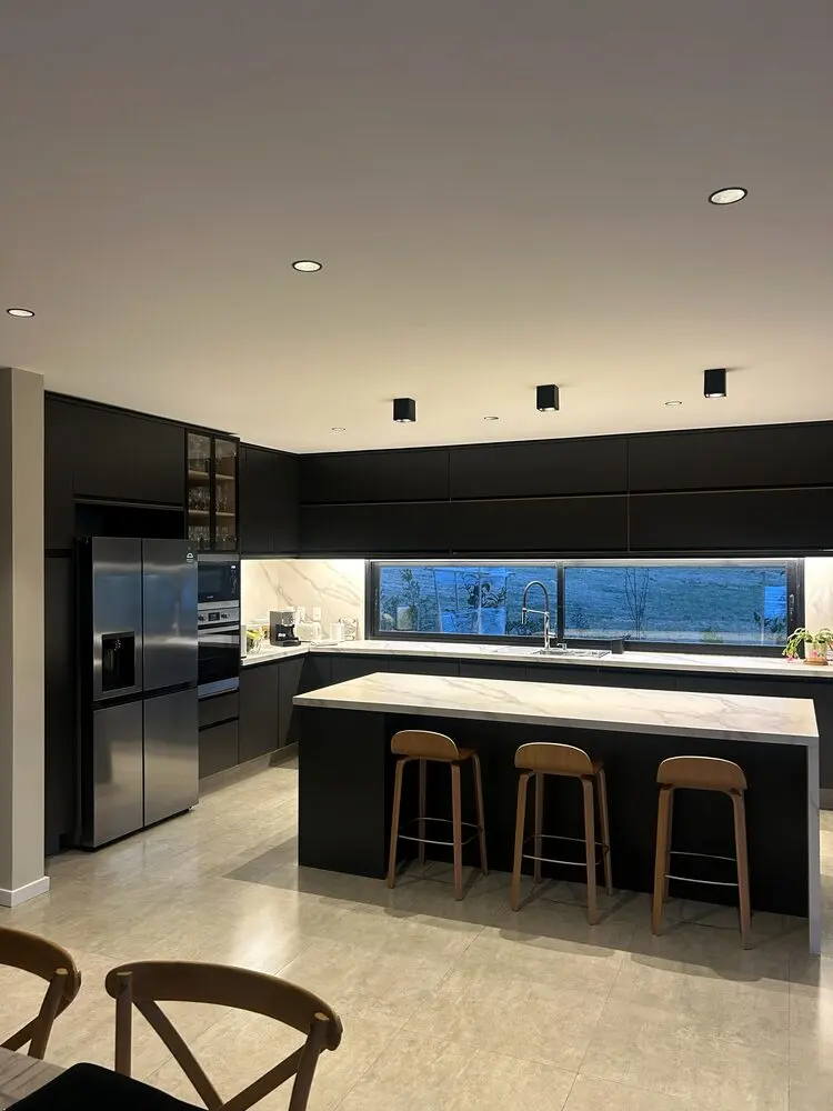 Cocina negra con isla de mesada blanca y banquetas de madera, iluminada de noche
